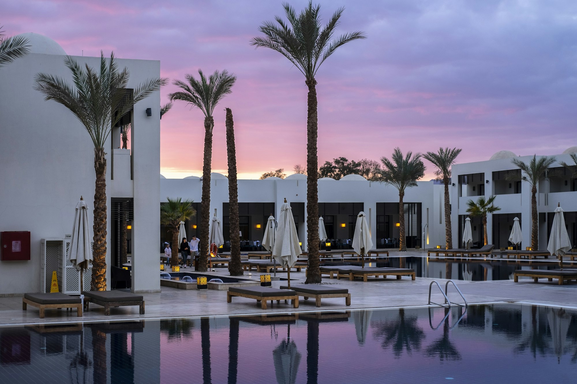Weiße Hotelanlage in der Abenddämmerung mit Palmen und einem Pool, in dem sich der Himmel spiegelt.